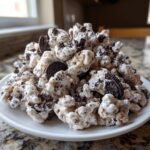A mound of white chocolate-coated cookies and cream puppy chow mixed with crushed Oreos on a white plate.