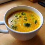 Close-up of creamy, vibrant yellow broccoli cheddar soup topped with florets in a white mug.