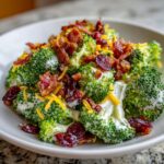 Close-up of creamy broccoli salad featuring fresh broccoli florets, bacon bits, dried cranberries, and shredded cheddar cheese.