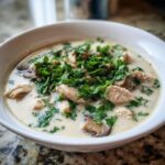 A white bowl filled with creamy coconut soup featuring chunks of chicken, sliced mushrooms, and topped generously with fresh green cilantro or parsley.