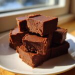 A stack of rich, dark chocolate fudge squares piled high on a white plate, catching bright sunlight.