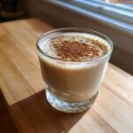 A glass of creamy coquito recipe topped generously with ground cinnamon, sitting on a wooden surface.