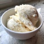 Two scoops of creamy, pale yellow cottage cheese ice cream served in a small white bowl.