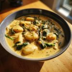 Close-up of a bowl of creamy, yellow gnocchi soup featuring soft gnocchi and wilted spinach, seasoned with pepper.