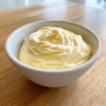A white bowl filled with thick, creamy, pale yellow homemade mayonnaise, resting on a wooden surface.