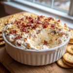 A creamy Neiman Marcus dip served in a white ramekin, topped generously with crumbled bacon and chopped nuts, surrounded by crackers.