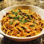 A white bowl filled with fettuccine noodles generously coated in a creamy peanut sauce and topped with fresh parsley.