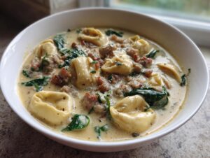 A close-up of a white bowl filled with creamy sausage tortellini soup, featuring pasta, crumbled sausage, and wilted spinach.