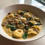 A white bowl filled with rich, creamy sausage tortellini soup, featuring cheese tortellini, crumbled sausage, and wilted spinach.