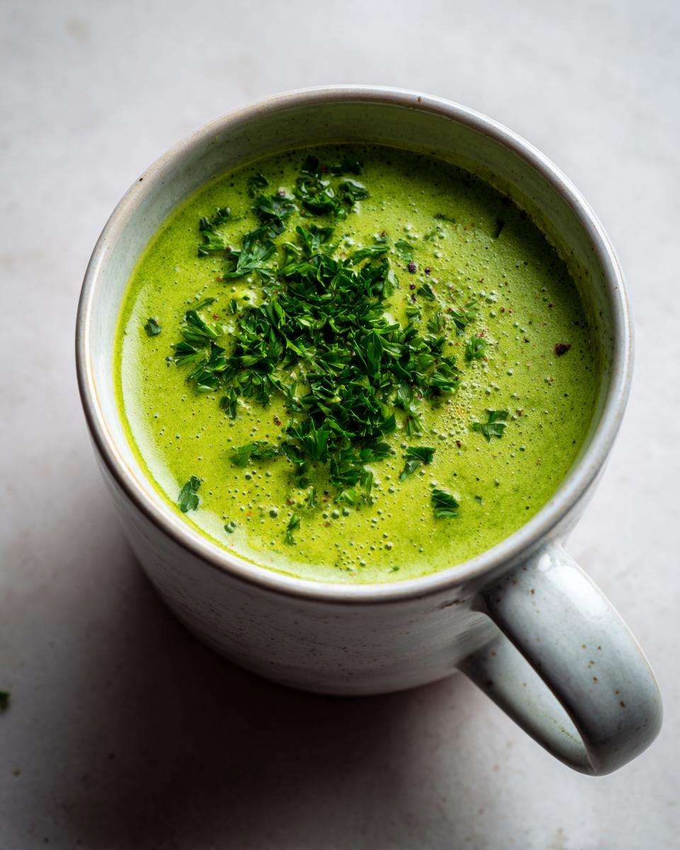 Overhead close-up of vibrant green creamy zucchini soup topped with fresh chopped parsley in a speckled ceramic mug.