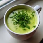Close-up of vibrant green creamy zucchini soup topped generously with fresh chopped parsley, served in a white mug.