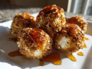 Four golden-brown, crispy fried goat cheese balls drizzled generously with amber honey on a white plate.