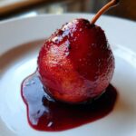 A single, whole poached pears, deeply stained red by wine syrup, resting in a pool of rich sauce on a white plate.
