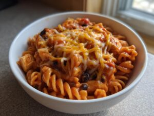 A white bowl filled with cheesy, saucy enchilada pasta featuring rotini noodles and black beans.