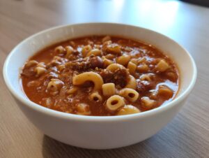 Close-up of a white bowl filled with rich, tomato-based pasta fagioli soup featuring elbow macaroni and ground meat.
