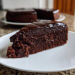 A decadent slice of flourless chocolate cake topped with shiny chocolate ganache, served on a white plate.