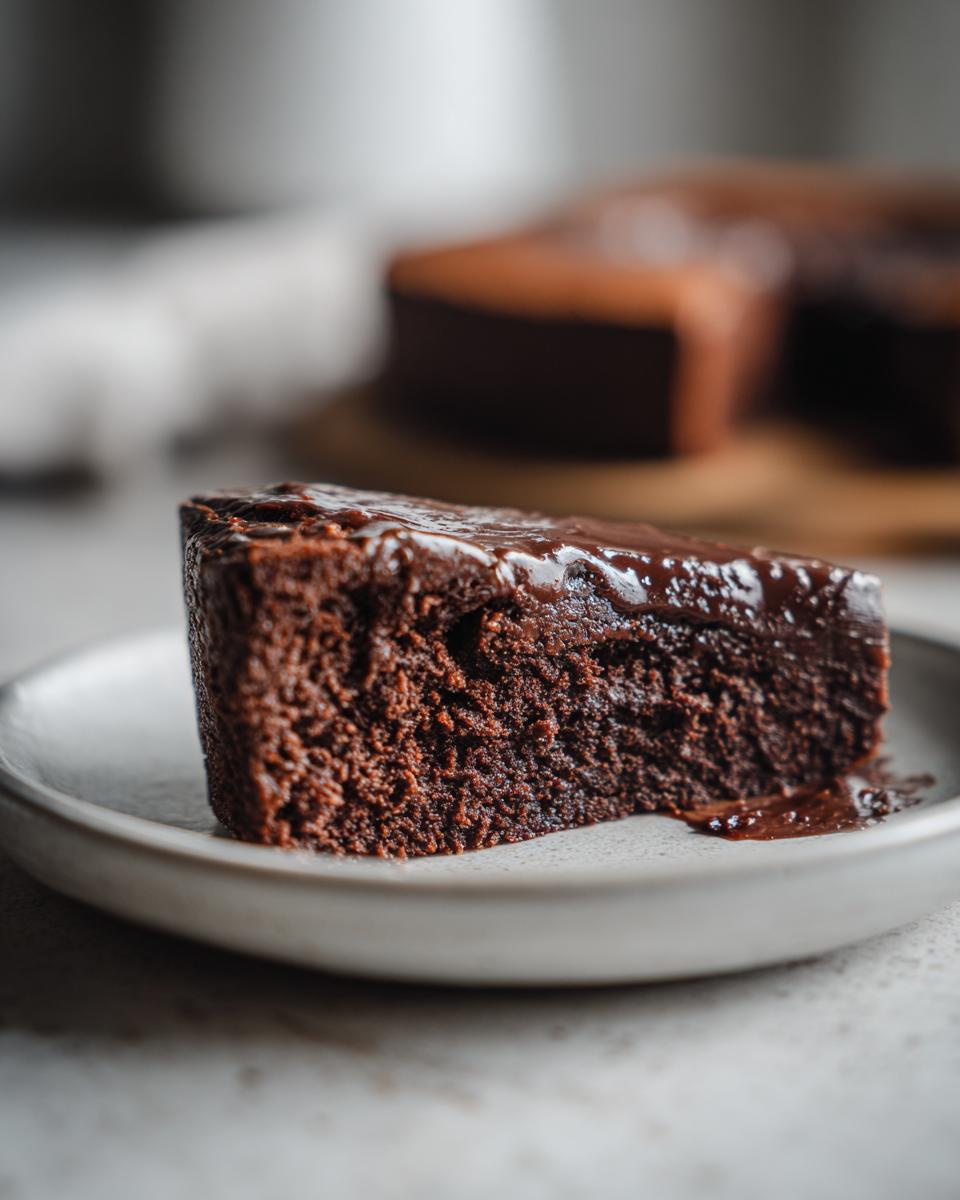 Close-up of a moist slice of flourless chocolate cake topped with shiny chocolate ganache on a light plate.