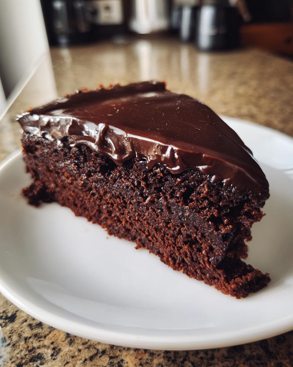 A close-up of a moist slice of flourless chocolate cake topped with glossy chocolate ganache on a white plate.