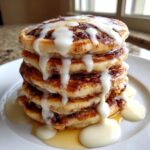 A tall stack of fluffy cinnamon roll pancakes drizzled heavily with white cream cheese icing.