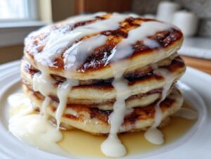 A tall stack of fluffy cinnamon roll pancakes drizzled heavily with white cream cheese icing.