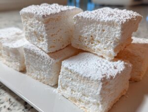 Close-up of several fluffy homemade marshmallows stacked on a white platter, heavily dusted with powdered sugar.