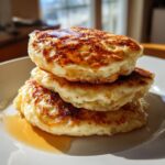 A close-up of a fluffy stack of three golden-brown sourdough pancakes drizzled with syrup.