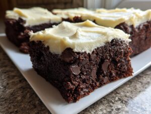 Close-up of a rich, dark gingerbread brownies slice topped with thick cream cheese frosting and visible chocolate chips.