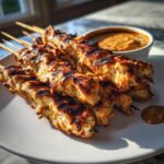 Close-up of perfectly grilled chicken satay skewers piled on a white plate next to a bowl of peanut dipping sauce.