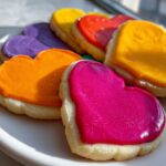 Close-up of heart-shaped sugar cookies topped with vibrant, shiny sugar cookie icing that hardens in pink, orange, yellow, and purple.