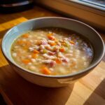 A close-up of a warm bowl of navy bean soup, featuring white beans, diced carrots, celery, and ham pieces.