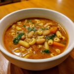 Close-up of a white bowl filled with rich, tomato-based pasta fagioli soup, featuring beans, carrots, and ditalini pasta.
