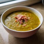 A close-up of a thick, green pea soup topped with shredded ham, served in a white bowl.