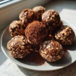 A close-up of several homemade protein balls coated in oats and cocoa powder, resting on a light gray plate.
