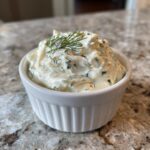 A close-up of creamy, homemade tartar sauce recipe garnished with fresh dill in a small white bowl.
