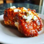A close-up of a piece of hot honey feta chicken, glazed in a bright orange sauce and topped with crumbled feta cheese.