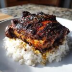 A close-up of perfectly cooked Jamaican jerk chicken with a dark, spicy crust resting on a bed of fluffy white rice.