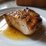 Close-up of a perfectly cooked, juicy baked chicken breast piece resting in its own pan juices, topped with herbs.