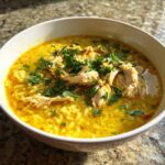 A close-up of a bowl of vibrant yellow Lemon Ginger Turmeric Chicken and Rice Soup, topped with shredded chicken and fresh parsley.