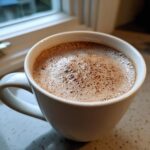 Close-up of a white mug filled with frothy mexican hot chocolate, set near a window.
