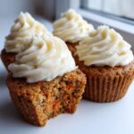 Close-up of moist carrot cake muffins topped with swirl of cream cheese frosting; one muffin has a bite taken out showing shredded carrots.