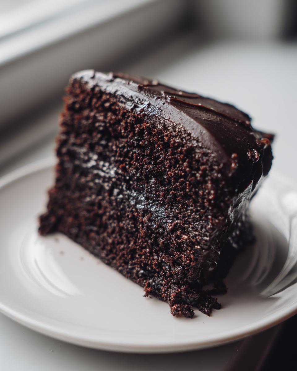 A close-up of a rich slice of moist chocolate cake topped with glossy dark chocolate frosting.