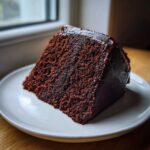A close-up of a rich, dark slice of moist chocolate cake topped with glossy chocolate frosting on a white plate.
