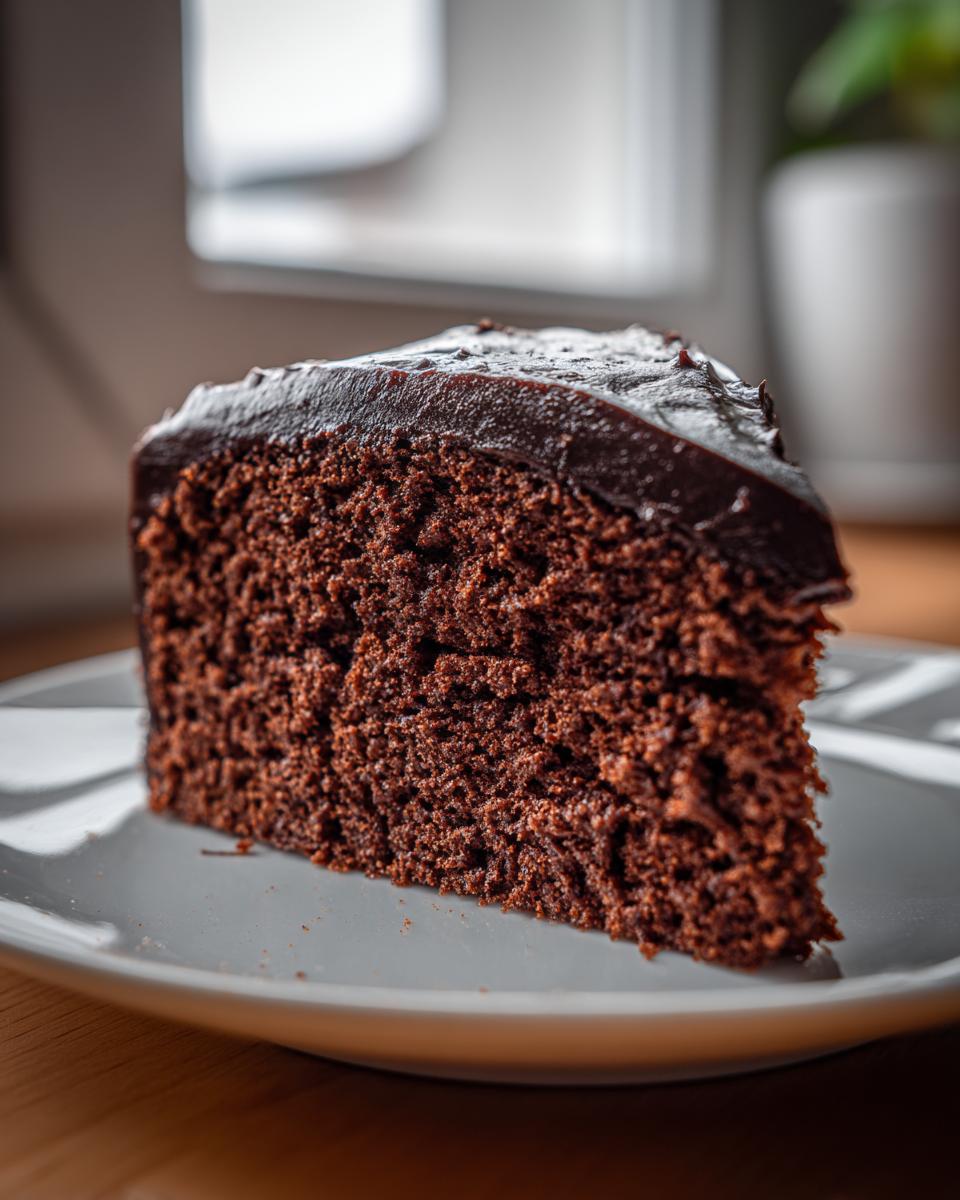 Close-up of a thick slice of moist chocolate cake topped with glossy dark chocolate frosting.