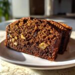 Two moist slices of dark brown date nut bread showing visible walnuts on a white plate.
