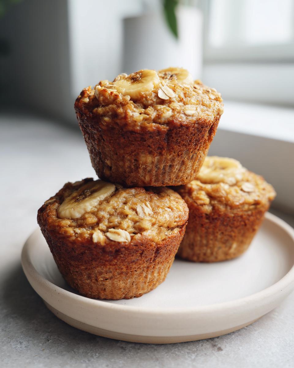 Three moist oatmeal muffins stacked on a small plate, topped with banana slices and oats.