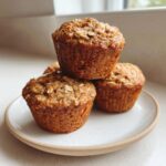 A stack of three golden brown, moist oatmeal muffins topped with visible oats, resting on a small ceramic plate.