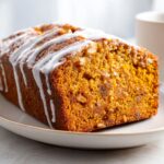A loaf of rich, orange-colored moist pumpkin bread topped with thick white icing drizzled across the top.