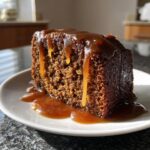 A rich, dark slice of sticky toffee pudding recipe covered in dripping caramel sauce on a white plate.