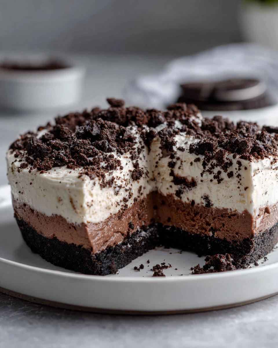 A cross-section view of a no bake Oreo pie showing a dark crust, chocolate mousse layer, white cream layer, and crumbled topping.