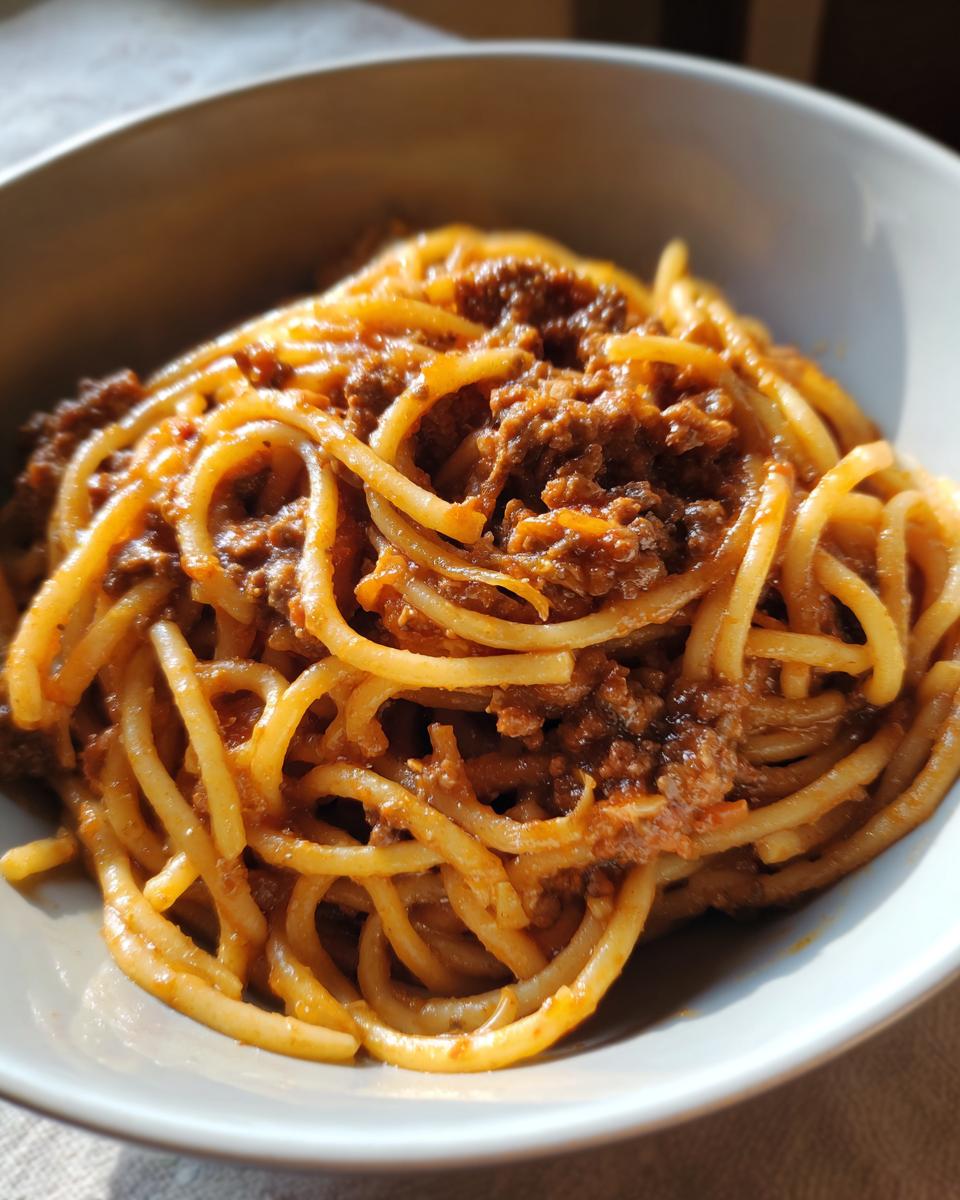 Close-up of thick spaghetti noodles generously coated in rich, slow-cooked bolognese sauce served in a light gray bowl.